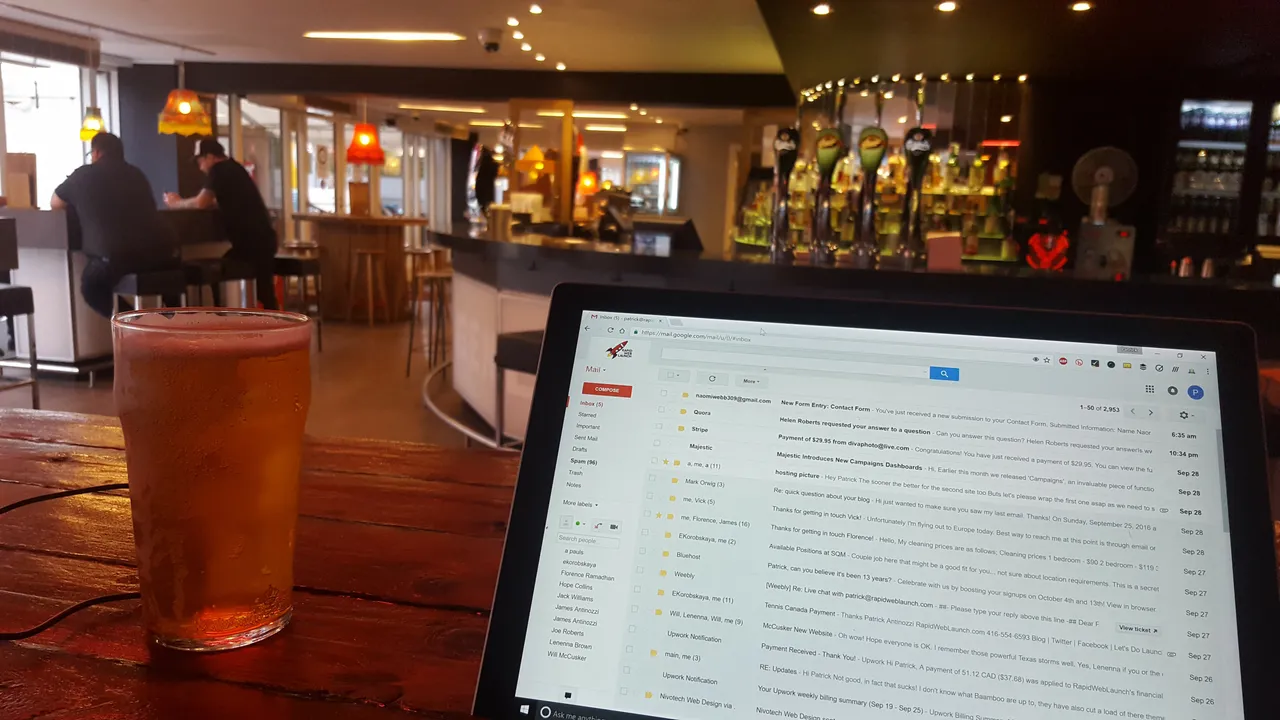 A laptop on a wooden bar table next to a glass of beer, with the pub's bar and taps in the background