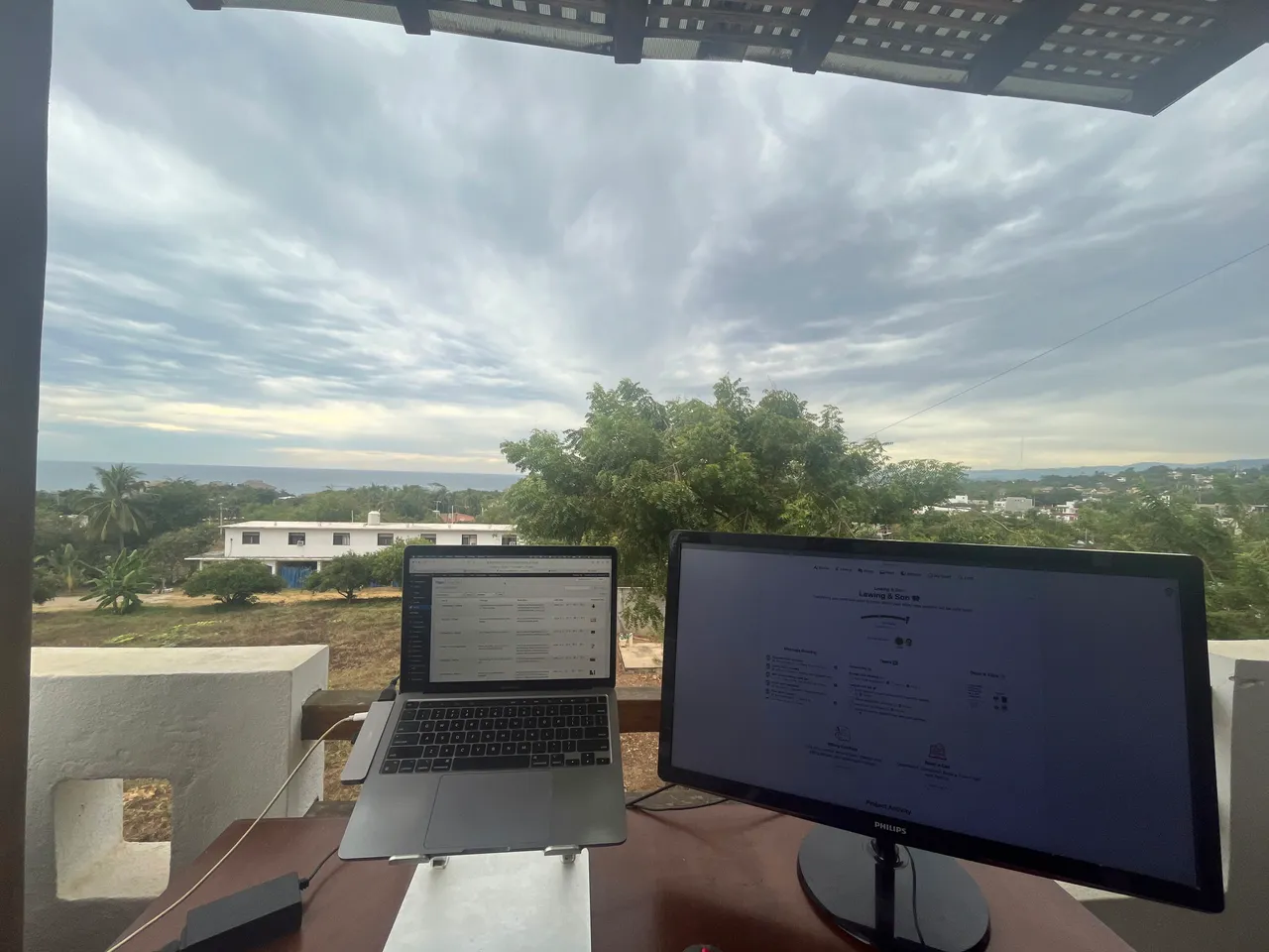 An outdoor workspace with a laptop and a monitor on a desk, set against a green landscape and a cloudy sky